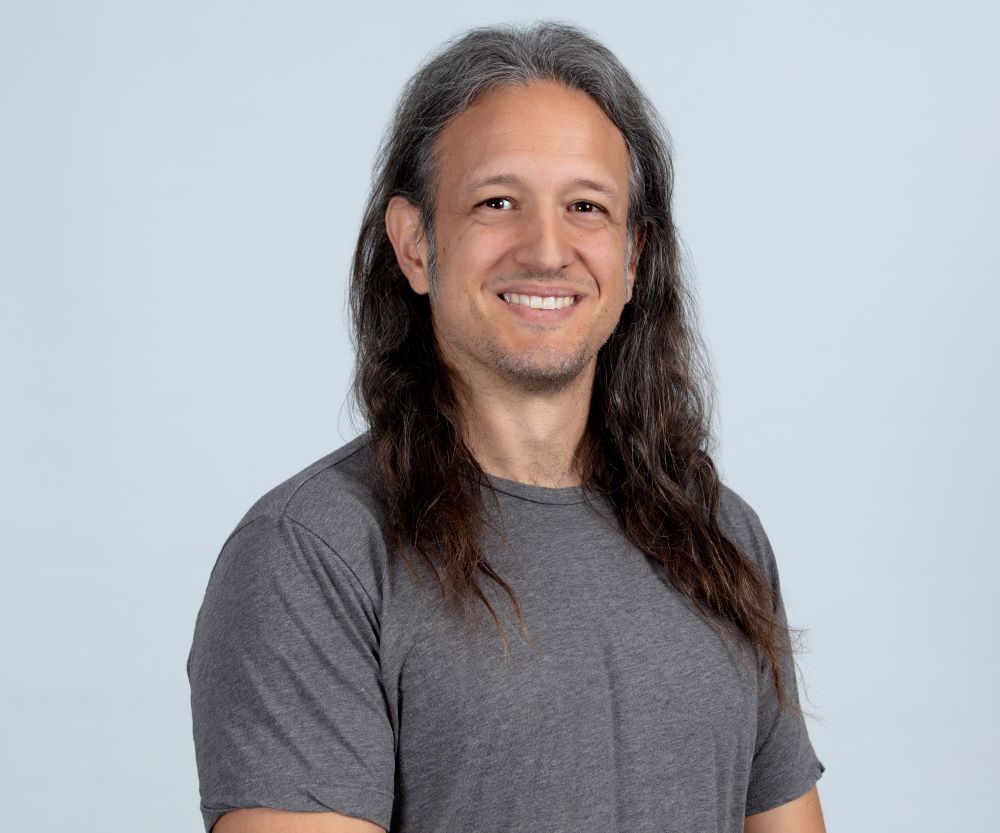 A professional man with long hair, smiling, wearing a gray t-shirt against a light background.