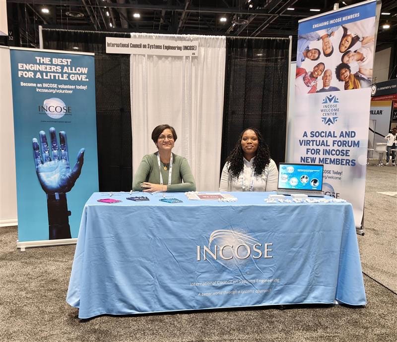 Junior woman and woman with long braids seated at INCOSE booth with banners at conference.