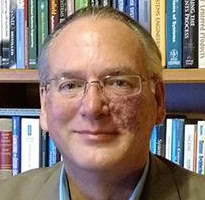 A professional man smiling in front of a bookshelf filled with books.