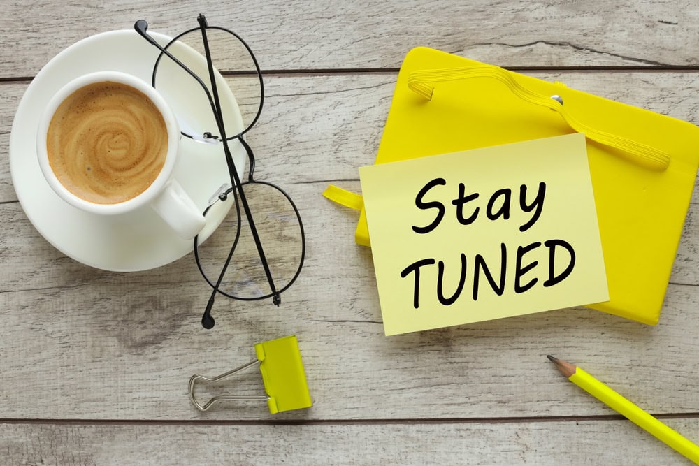 Coffee cup with eyeglasses on wooden table, "Stay TUNED" note, yellow folder, and pen.